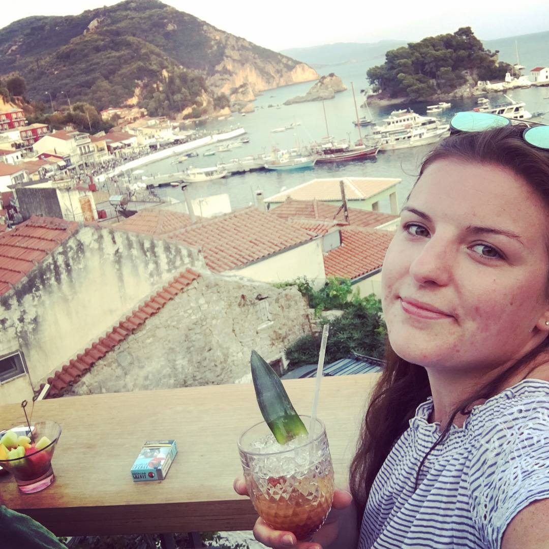 Woman holding a drink on a terrace overlooking a coastal village, harbour and boats with hills in the background image