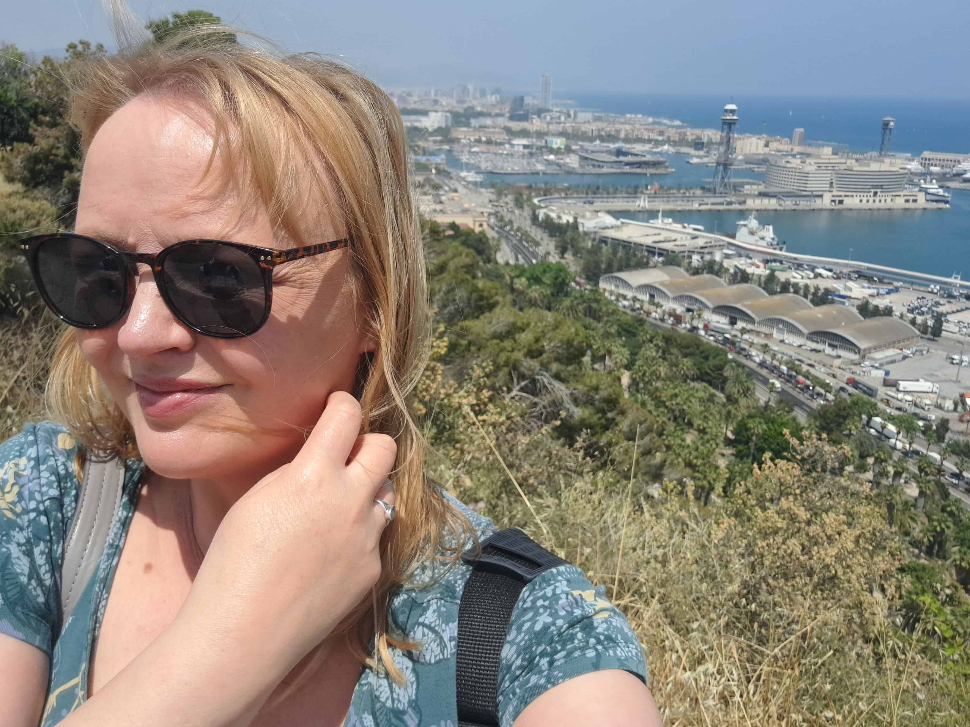Woman wearing sunglasses taking a selfie on a hillside overlooking a coastal city and harbour on a sunny day image