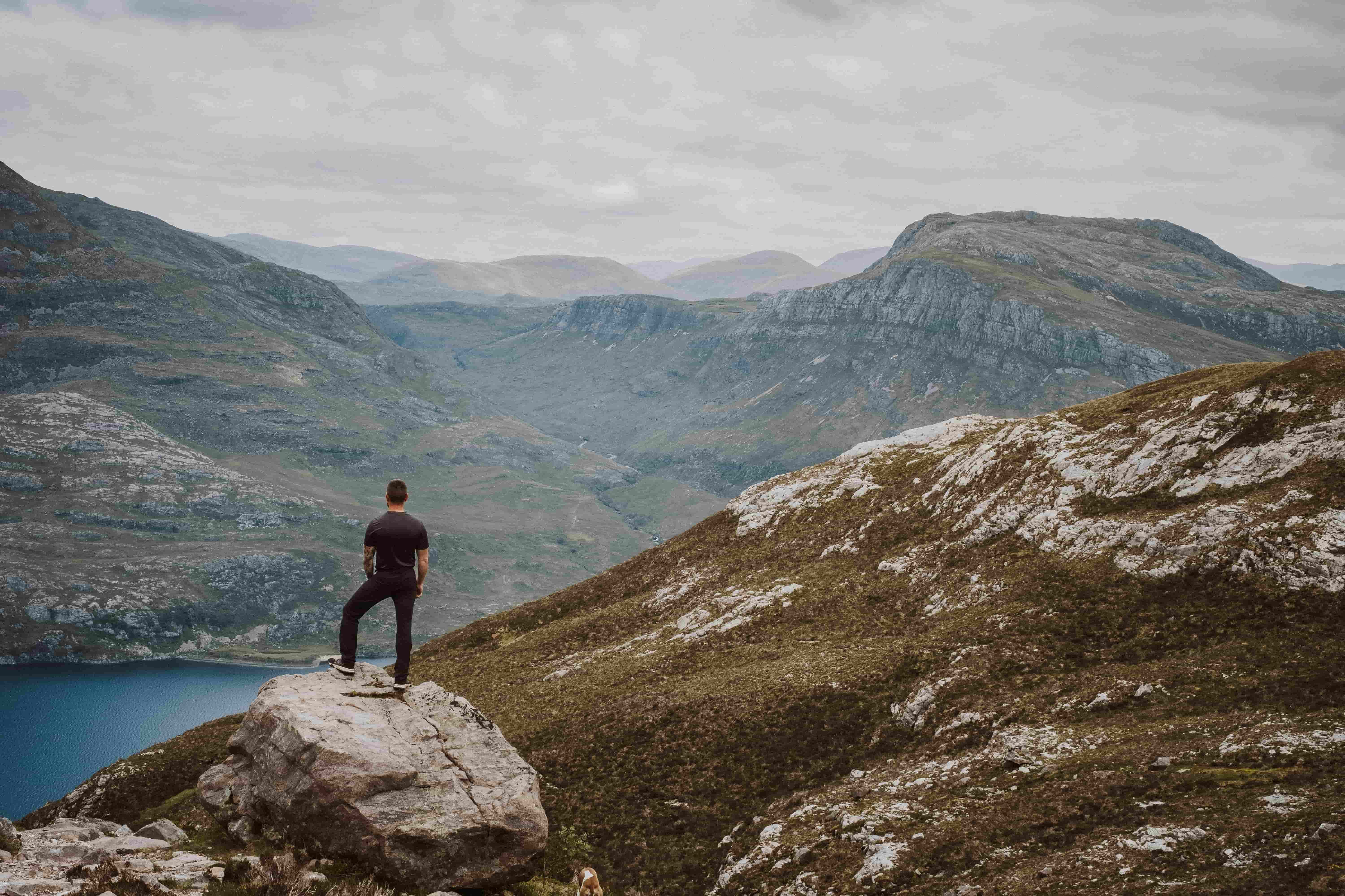 Person standing on a rocky outcrop overlooking a remote Scottish mountain valley and loch beneath an overcast sky. image