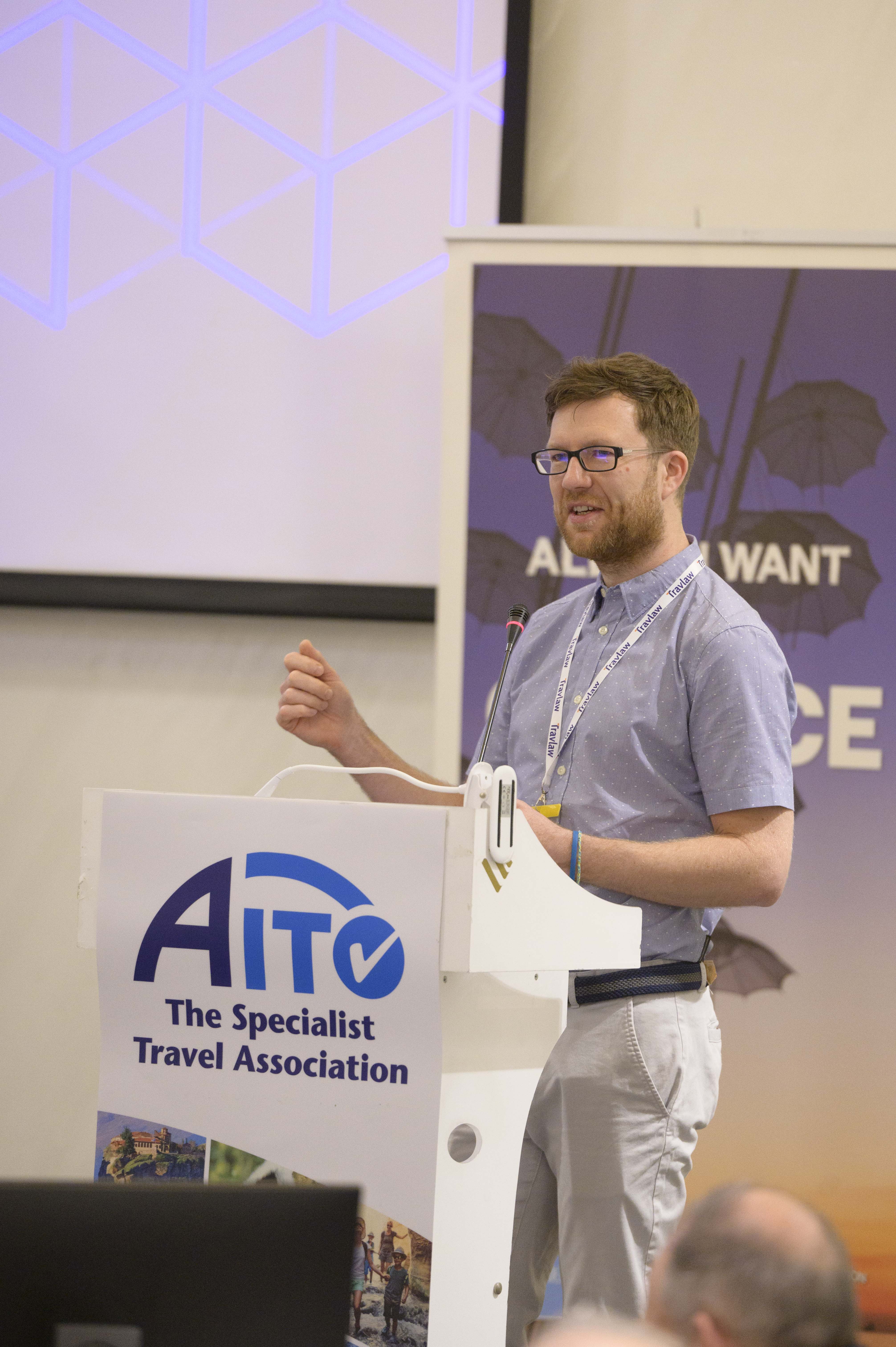 Speaker presenting at a podium branded with the AITO Specialist Travel Association logo during a travel industry event. image