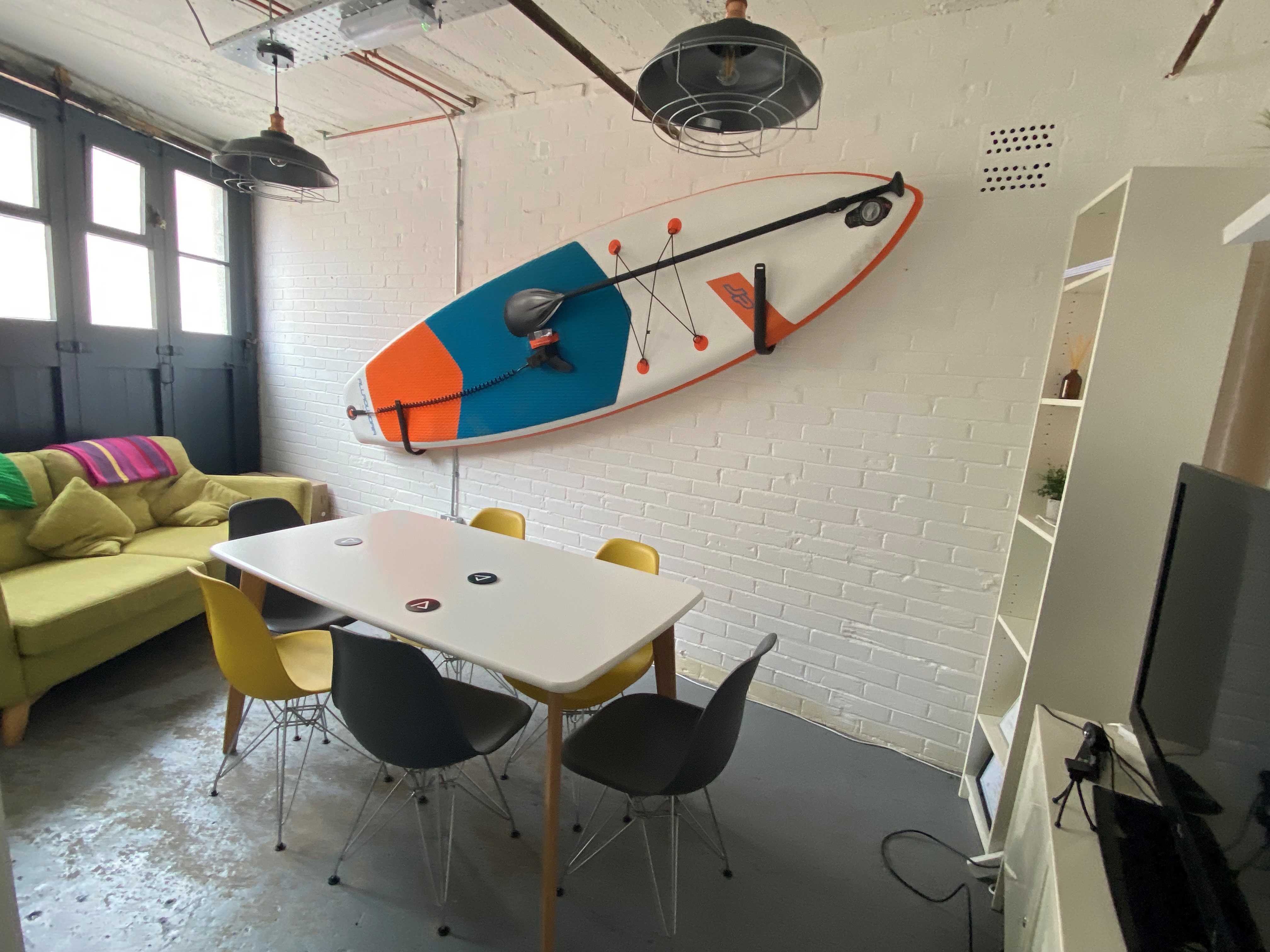 A small, funky meeting room with a white brick wall, a mounted paddleboard as decor, a rectangular table surrounded by mixed yellow and black chairs, and a green sofa beside large industrial-style windows. image