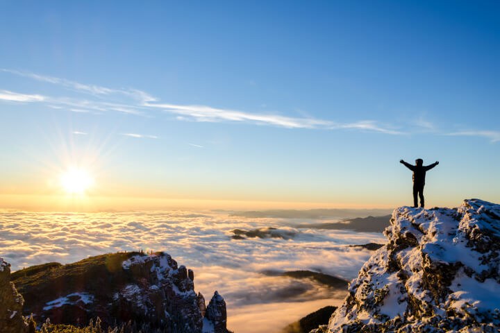A silhouette of a person standing on a snowy mountain peak with their arms outstretched toward a rising sun and a sea of clouds image